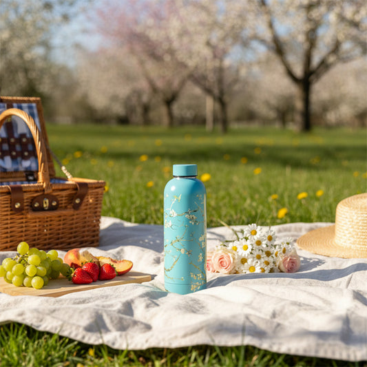 Vattenflaska med Van Goghs motiv av mandelblomster på en picknickfilt utomhus på våren, med mandelträd i full blom